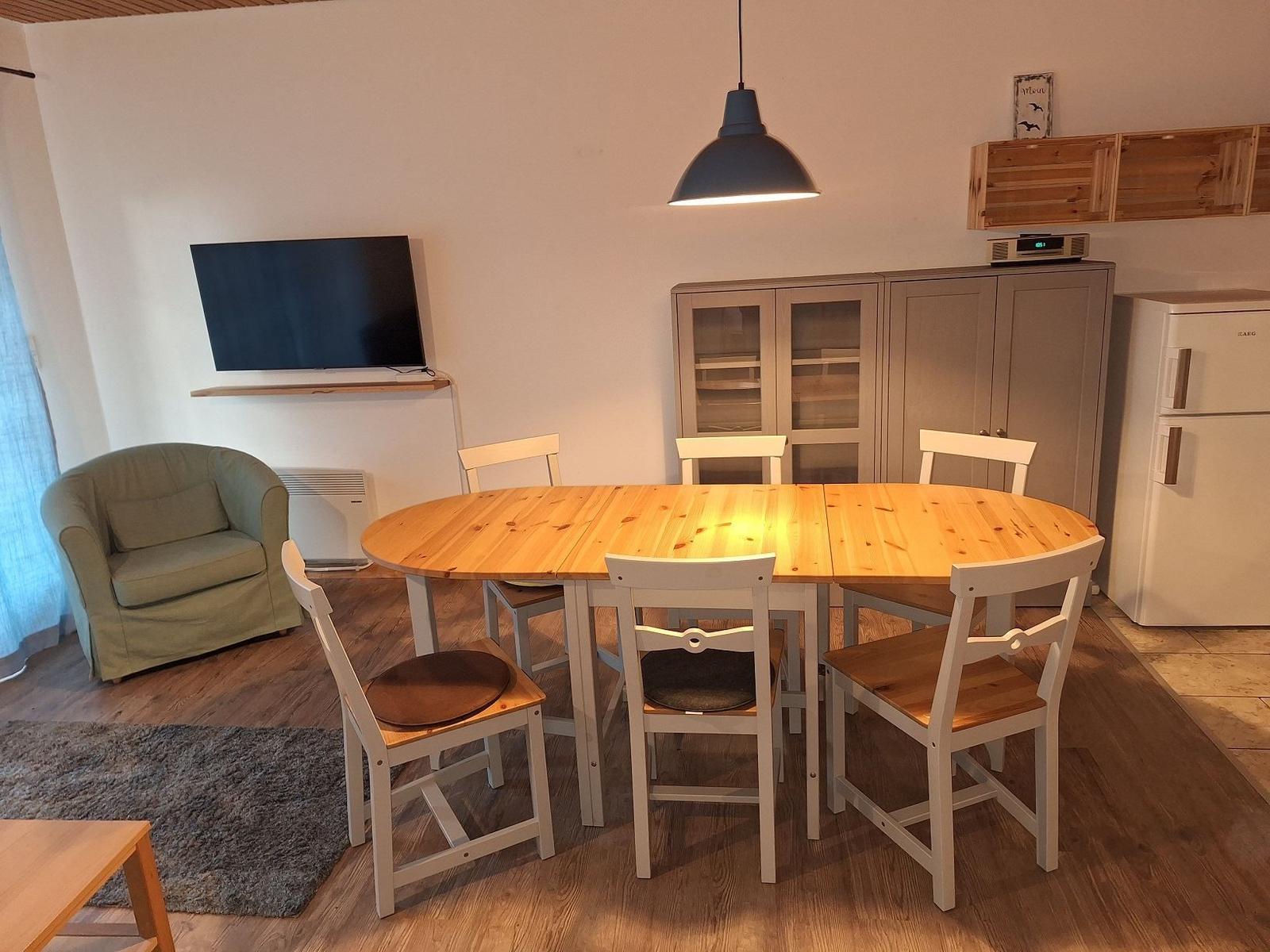 Essbereich mit Holztisch, weißen Stühlen und Fernseher an der Wand.