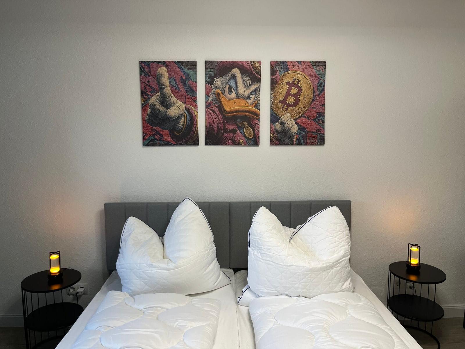 Zwei Betten mit weißen Kissen und Decken, Wandbilder mit Entenmotiv und Bitcoin, Nachttische mit Lampen.