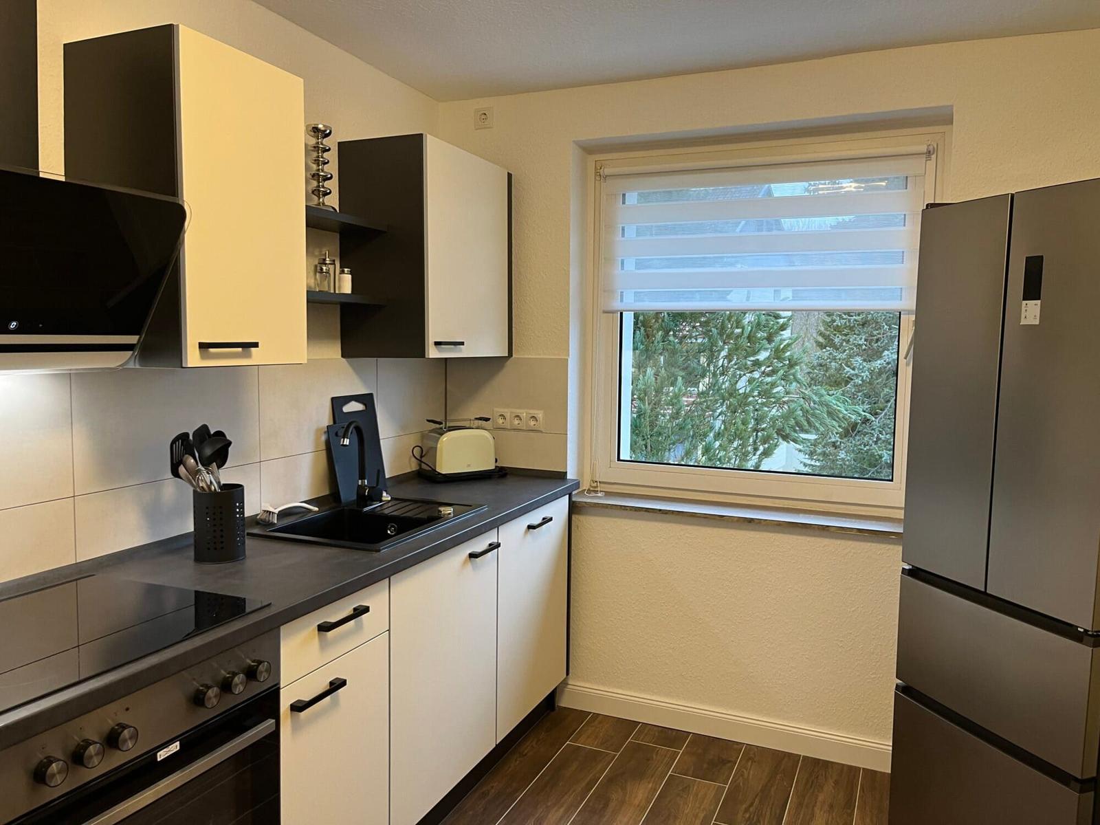 Küche mit Arbeitsplatte, Spüle, Herd und Kühlschrank. Fenster mit Jalousie und Blick auf Bäume.