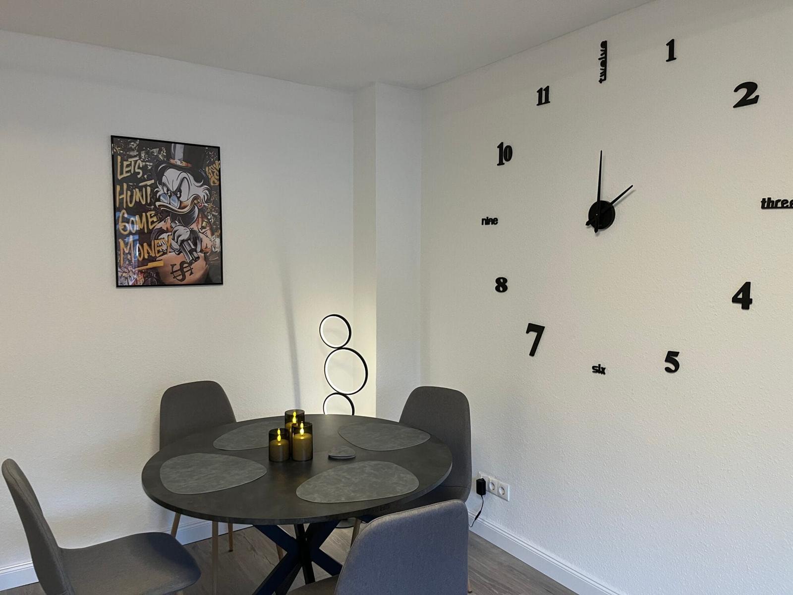 Runder Tisch mit Stühlen, Wanduhr mit Ziffern und Bild an der Wand.