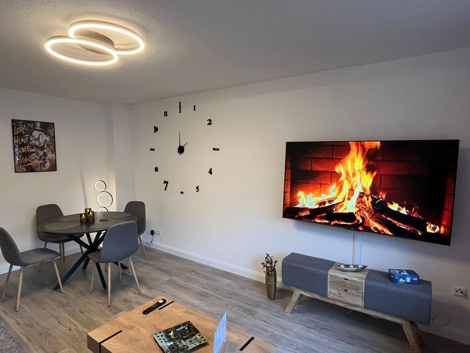 Wohnzimmer mit Esstisch, Wanduhr und Fernseher mit Kaminbild.