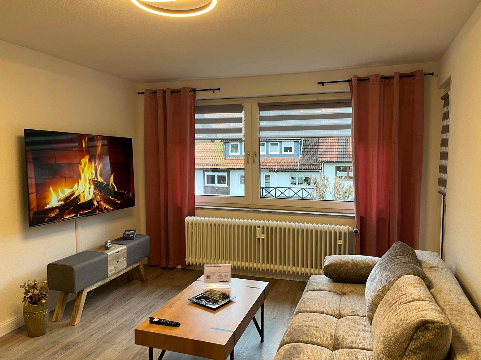 Wohnzimmer mit Sofa, Couchtisch und Fernseher an der Wand. Fenster mit Vorhängen und Heizkörper.