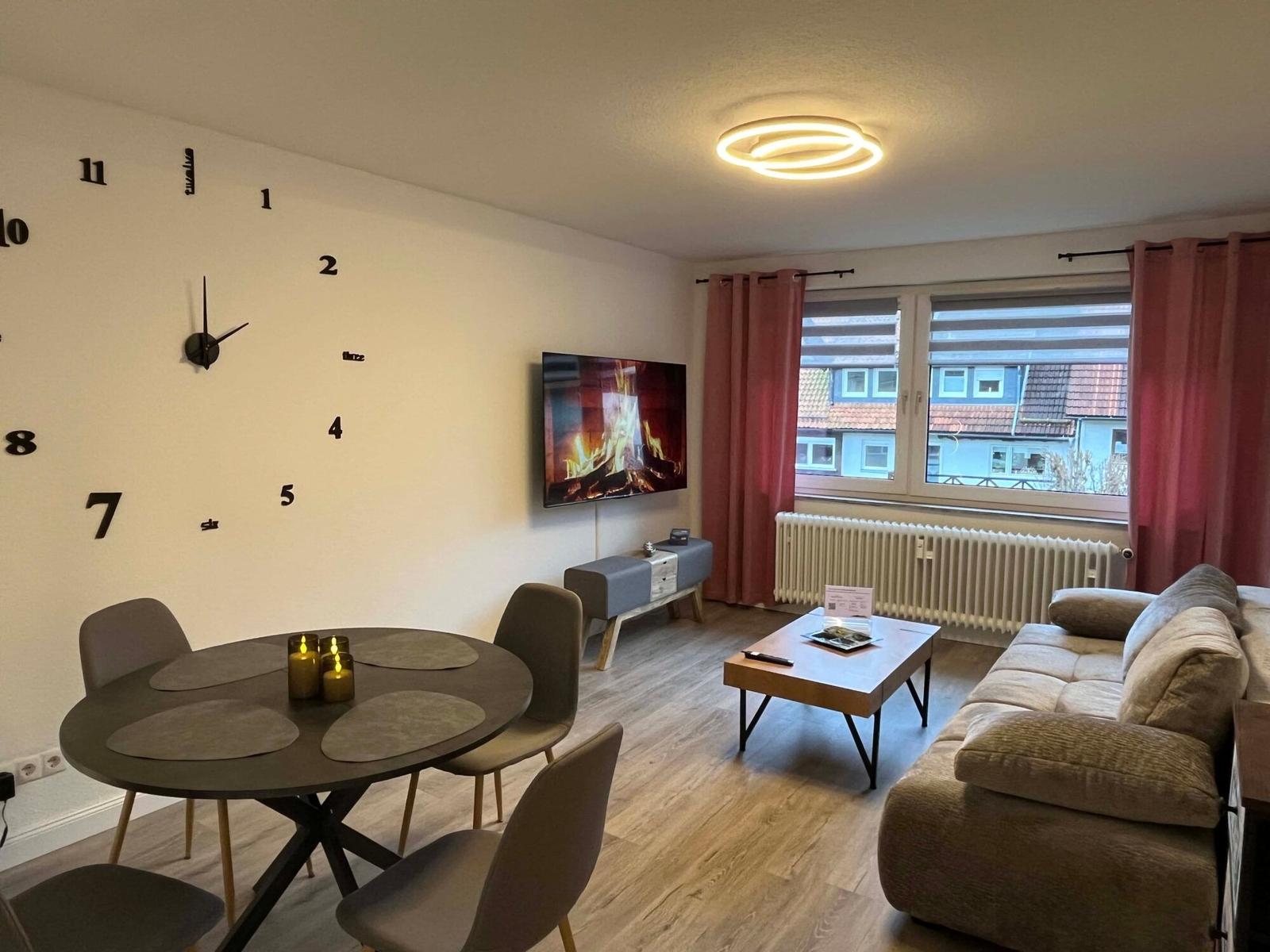 Wohn- und Essbereich mit Sofa, Esstisch und Wanduhr. Fenster mit Vorhängen.