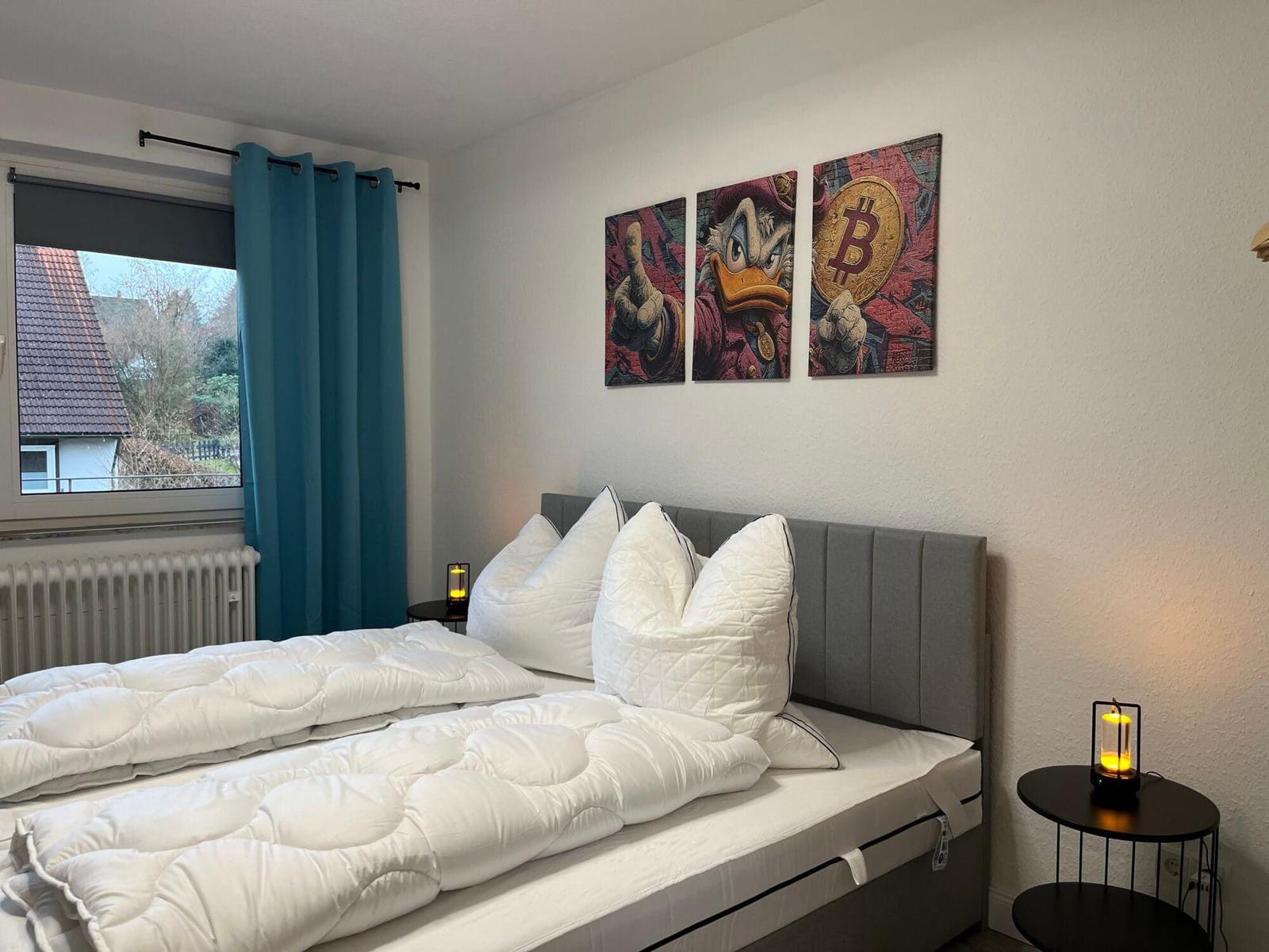 Schlafzimmer mit Bett, Wandbildern und Fenster mit Blick auf Dach und Bäume.