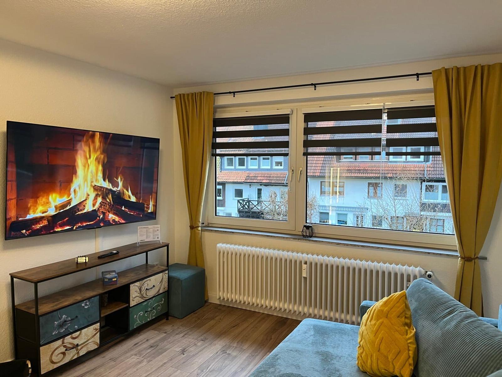 Wohnzimmer mit TV, Sofa und Fensterblick auf Gebäude.