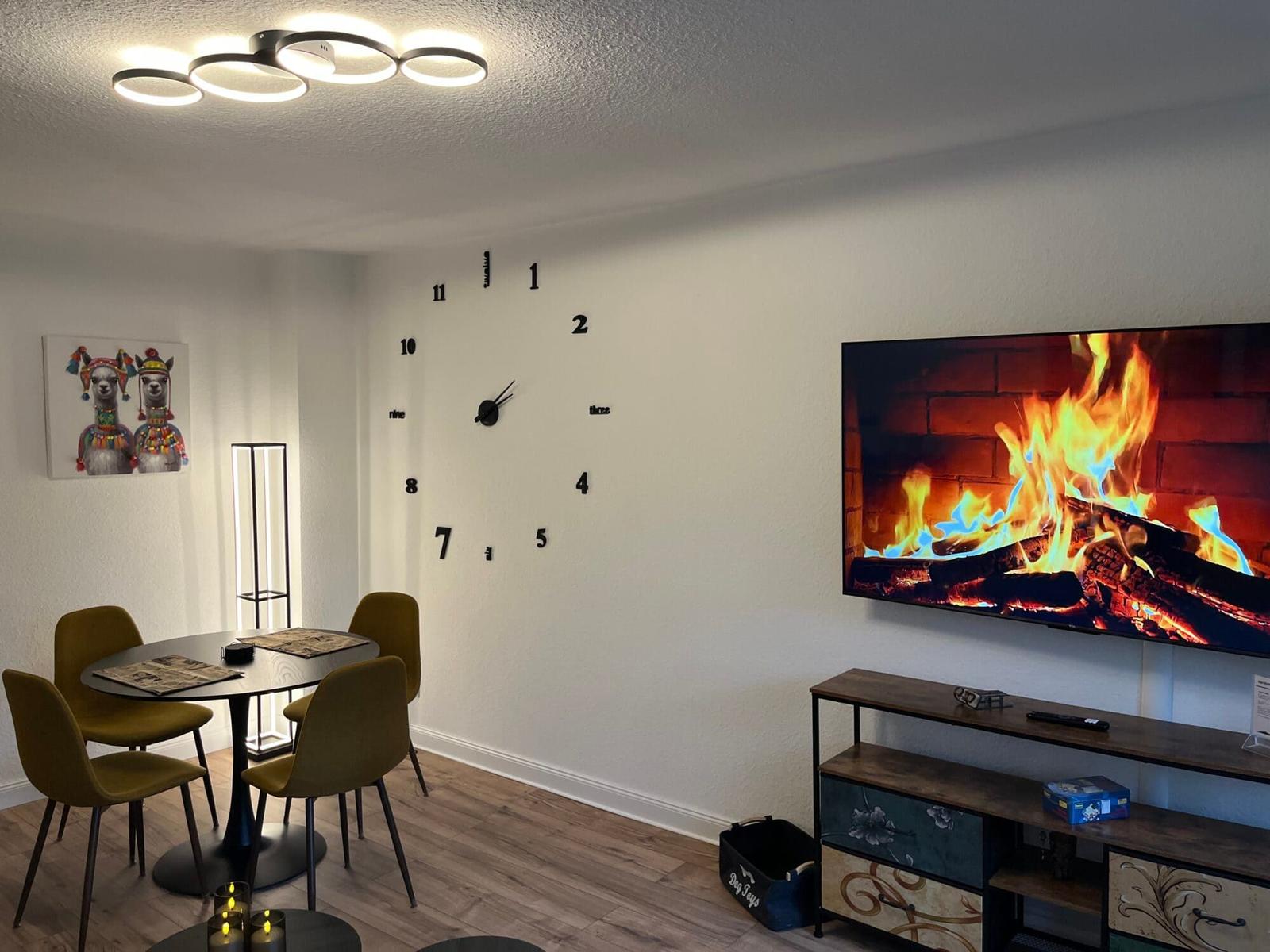 Wohn- und Essbereich mit Wanduhr, Fernseher und Tisch mit Stühlen.
