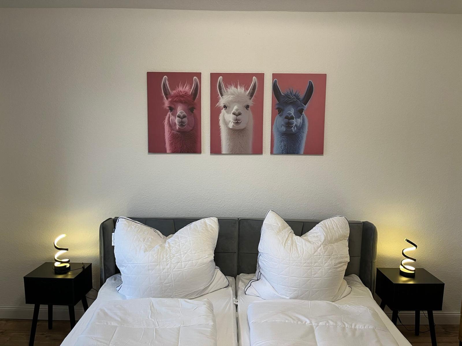 Zwei Betten mit weißen Kissen und Decken. An der Wand hängen drei Bilder von Alpakas.