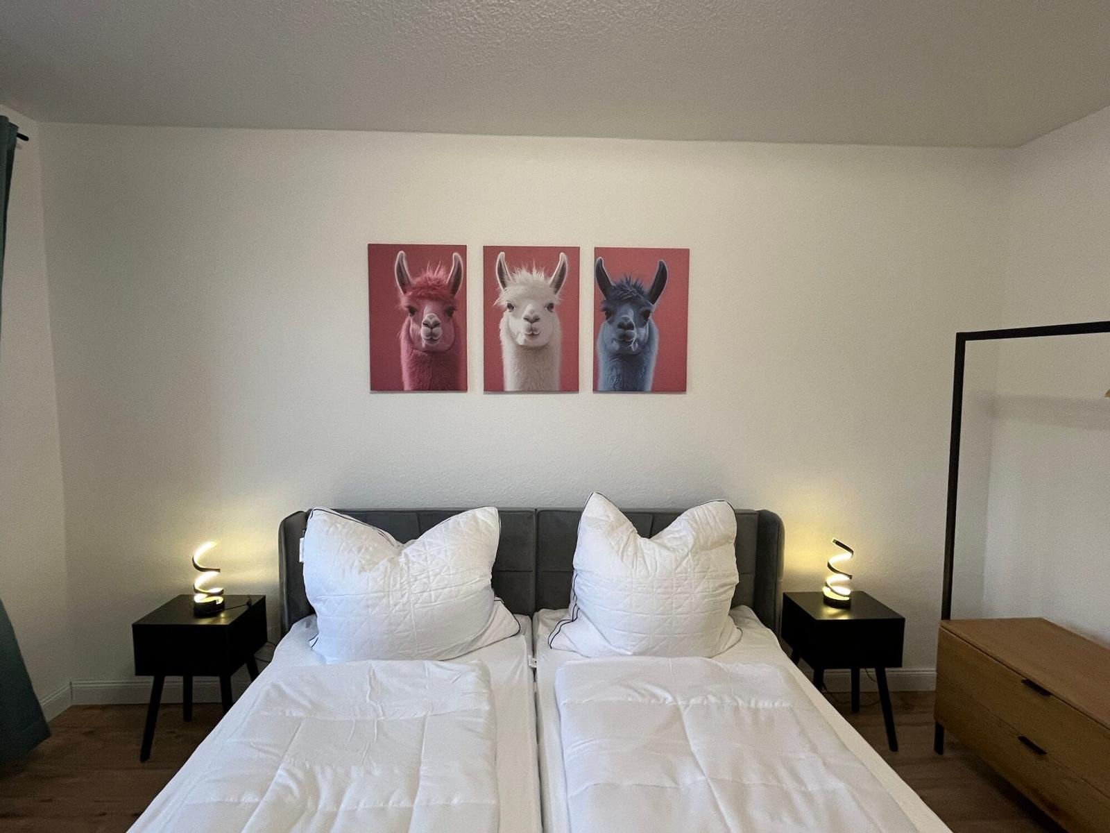 Zwei Betten mit weißen Bettwäschen, Wandbild mit drei Alpakas und Nachttische mit Lampen.