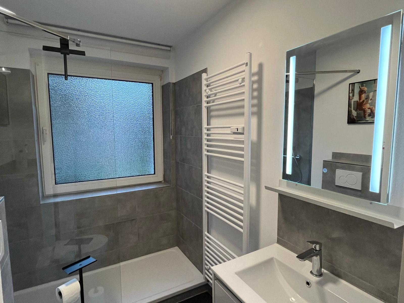 Badezimmer mit Dusche, Waschbecken und beleuchtetem Spiegel.