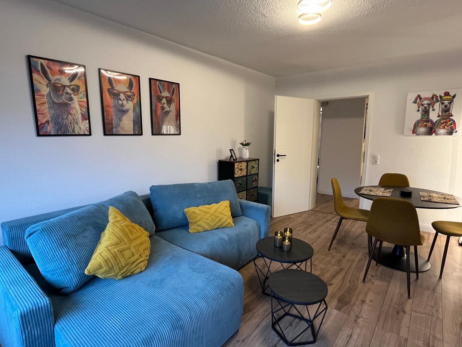 Blauer Sofa mit gelben Kissen, Essbereich mit Tisch und Stühlen, Wandbilder mit Lamas.