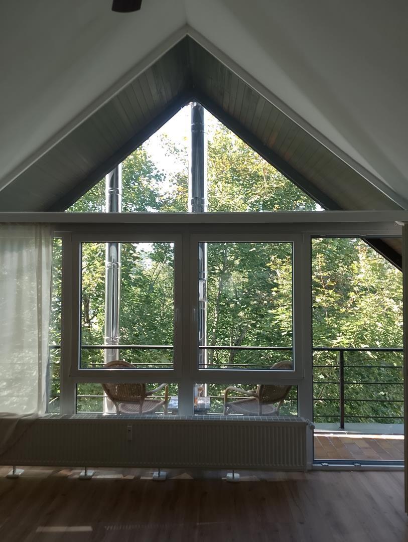 Large window overlooking balcony and forest. Wooden floor and gabled roof.