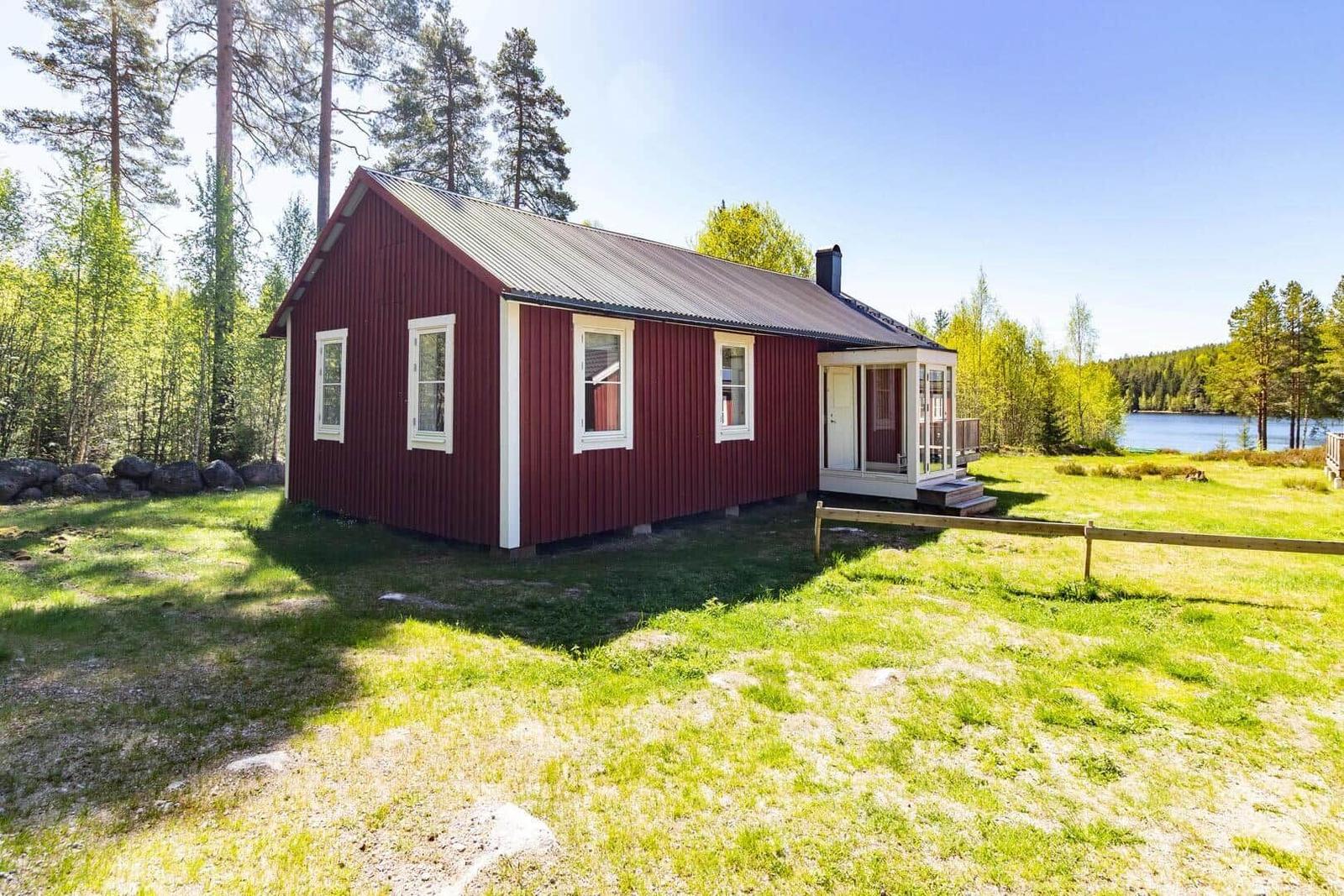 Rotes Holzhaus mit weißen Fenstern und Terrasse. Grasfläche und Wald im Hintergrund.