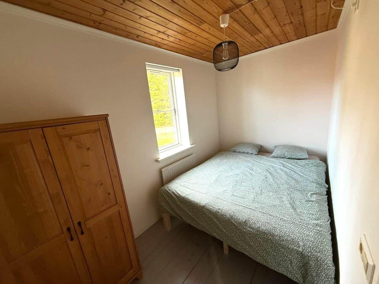 Schlafzimmer mit Bett, Kleiderschrank und Fenster. Holzdecke und Boden.