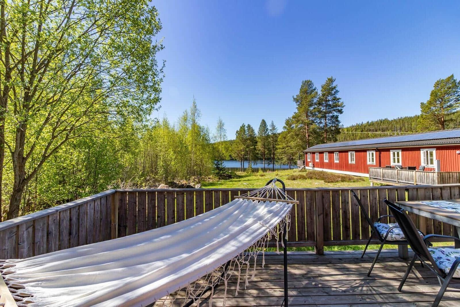 Terrasse mit Hängematte und Tisch, Blick auf roten Holzhaus und See.