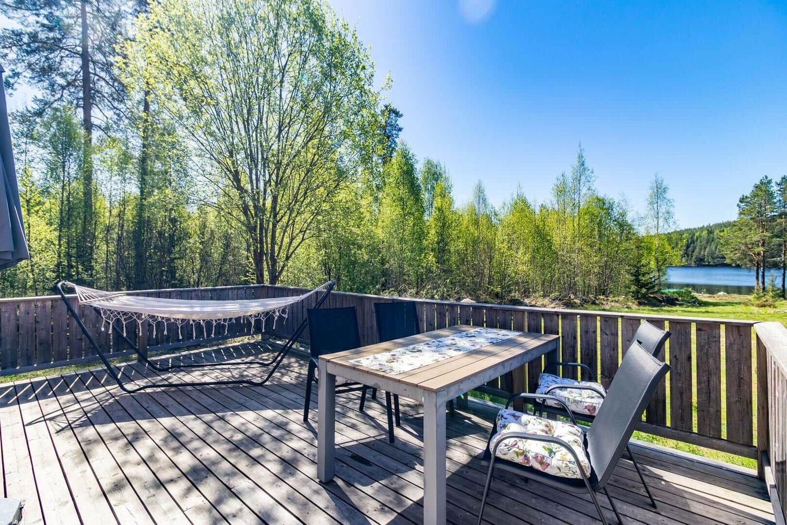 Terrasse mit Tisch, Stühlen und Hängematte. Blick auf Wald und See.