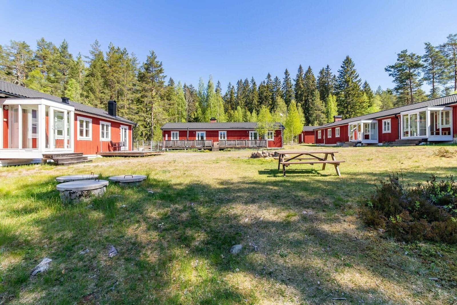 Rote Holzhäuser mit weißen Fensterrahmen stehen in einem grünen Grasfeld vor einem Wald.
