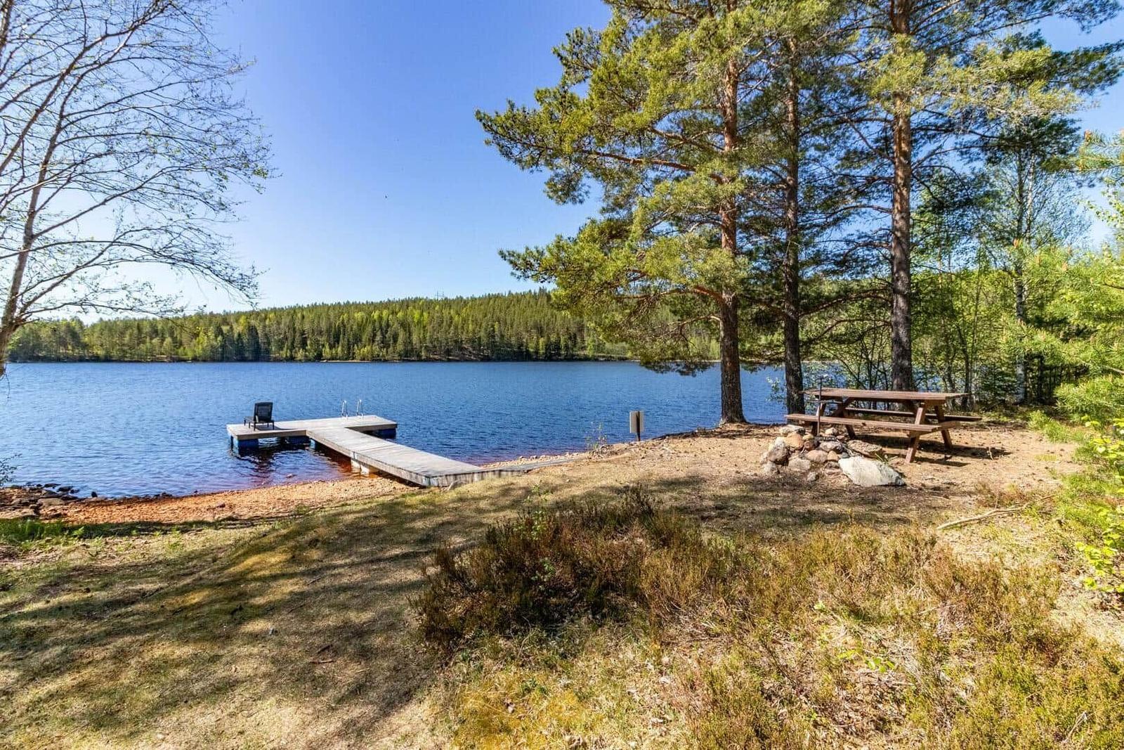 Holzsteg mit Liege am Seeufer unter Bäumen. Picknicktisch und Feuerstelle.