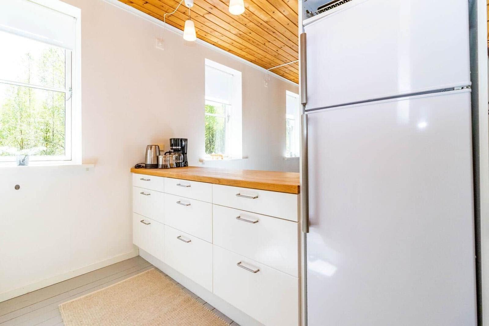 Küche mit weißen Schränken, Holzoberfläche und Kühlschrank. Fenster mit Blick auf Wald.