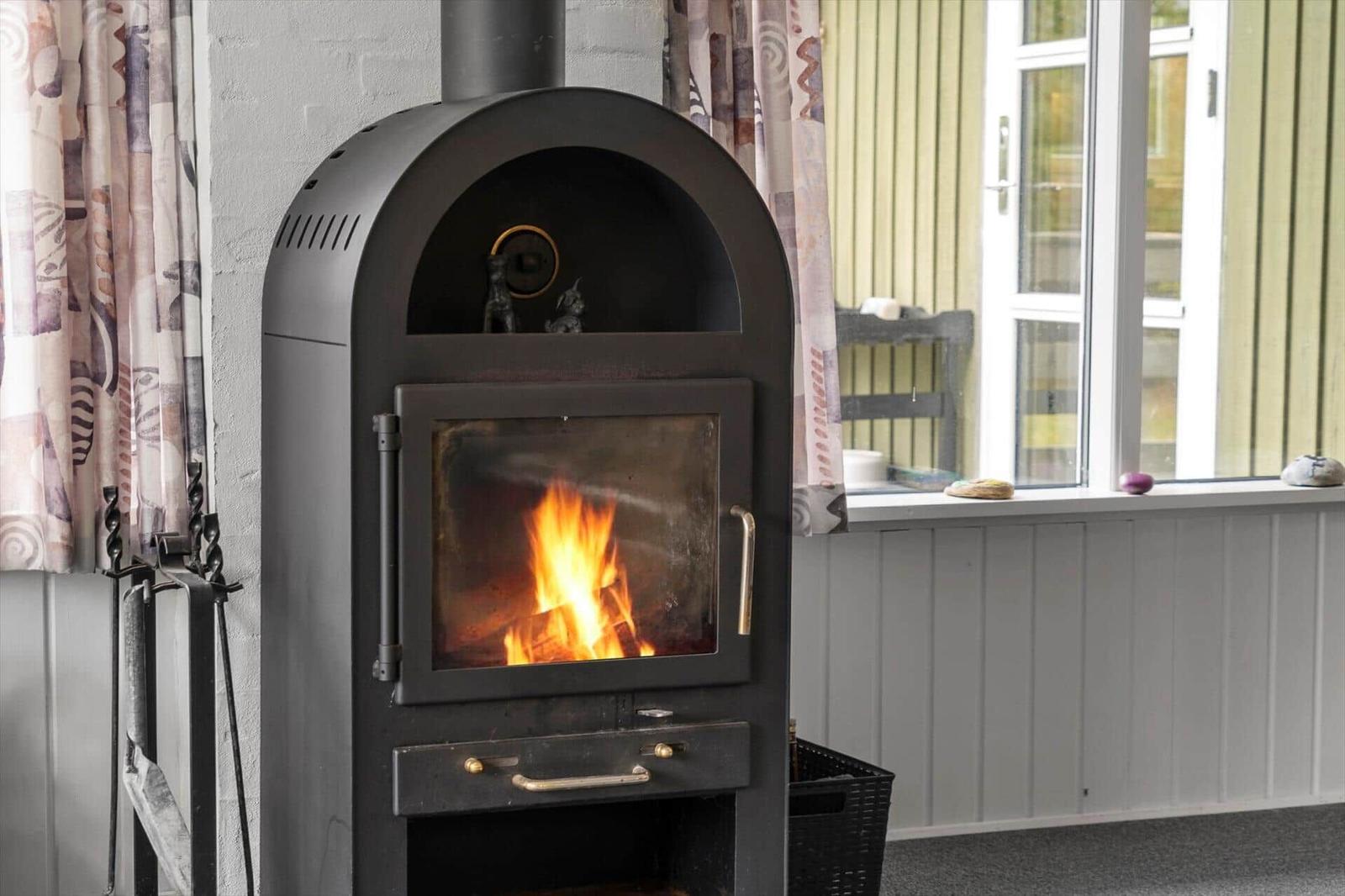 A wood-burning stove with a fire is positioned in front of a window with curtains.