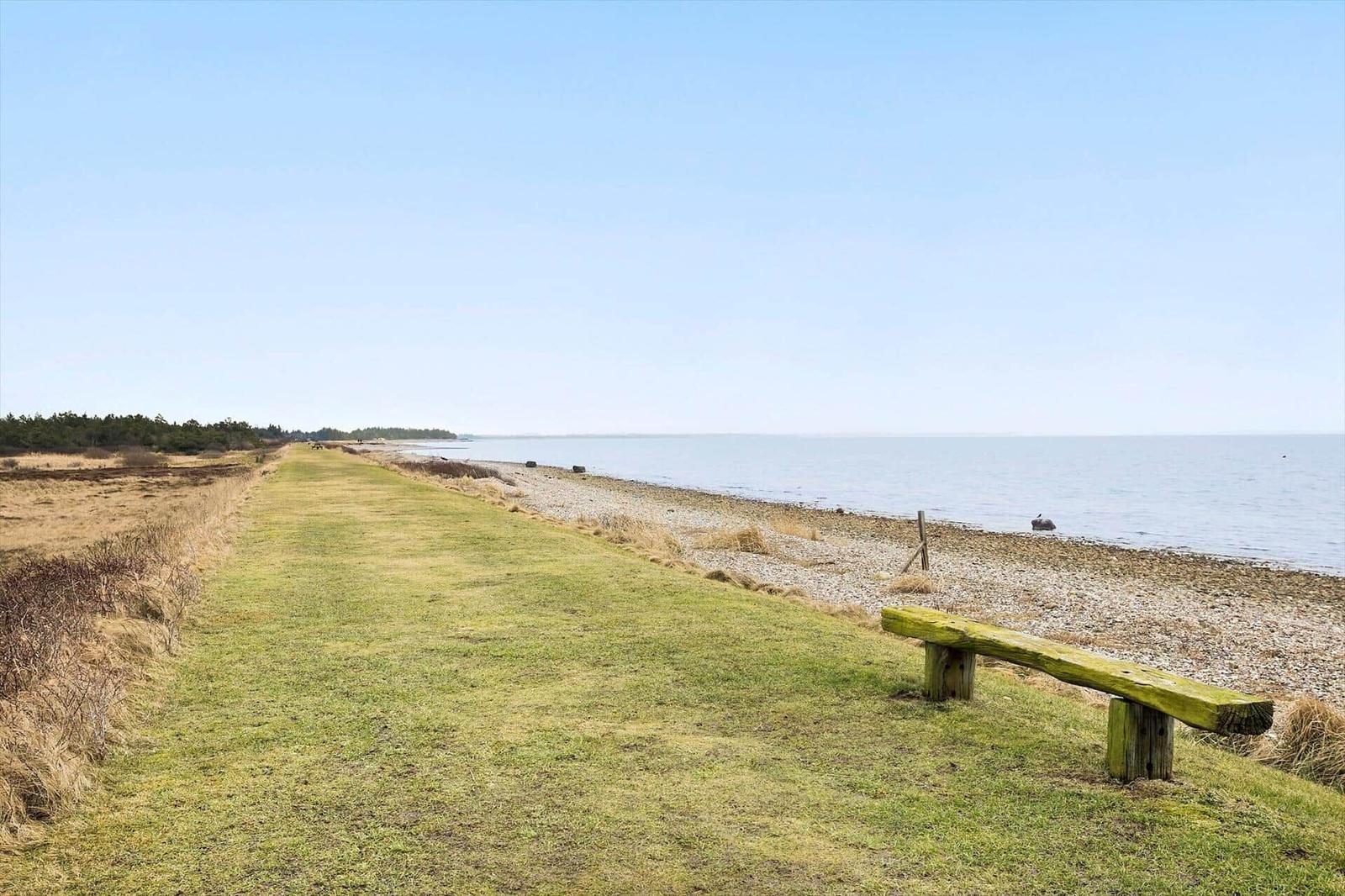 A grassy path runs alongside a pebbled beach to the sea. A wooden bench is in the foreground.