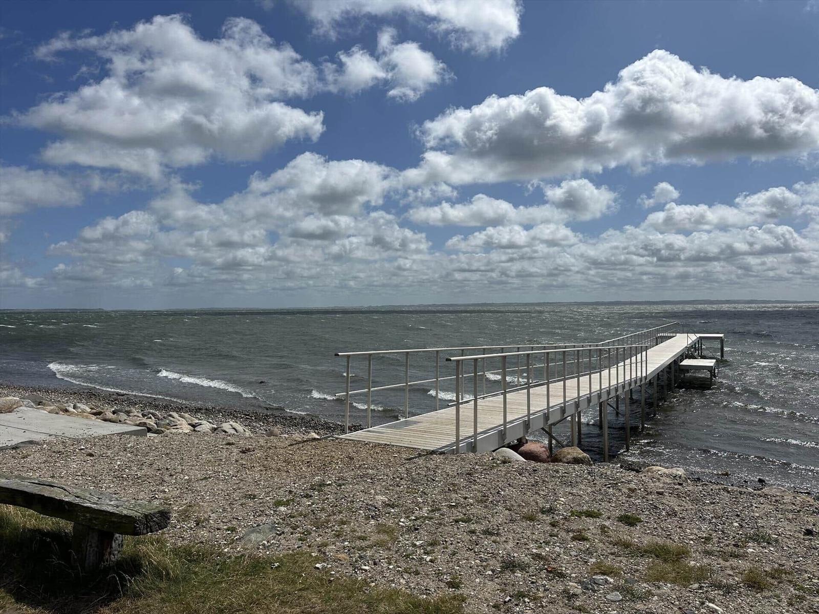 Wooden pier leads to the water. Rocky ground and sky with clouds.