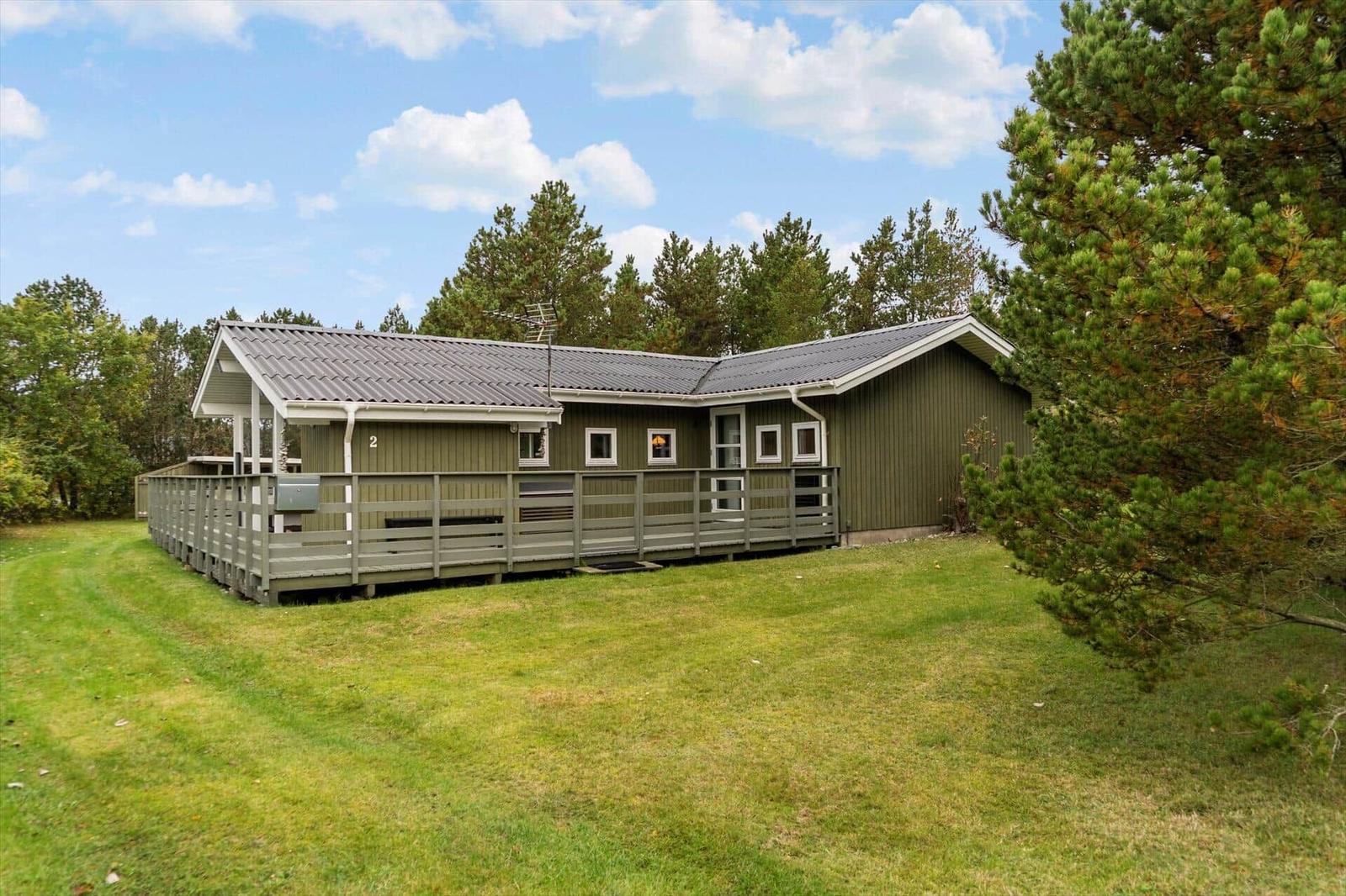 Green holiday home with balcony and terrace, surrounded by grass and trees under blue sky.