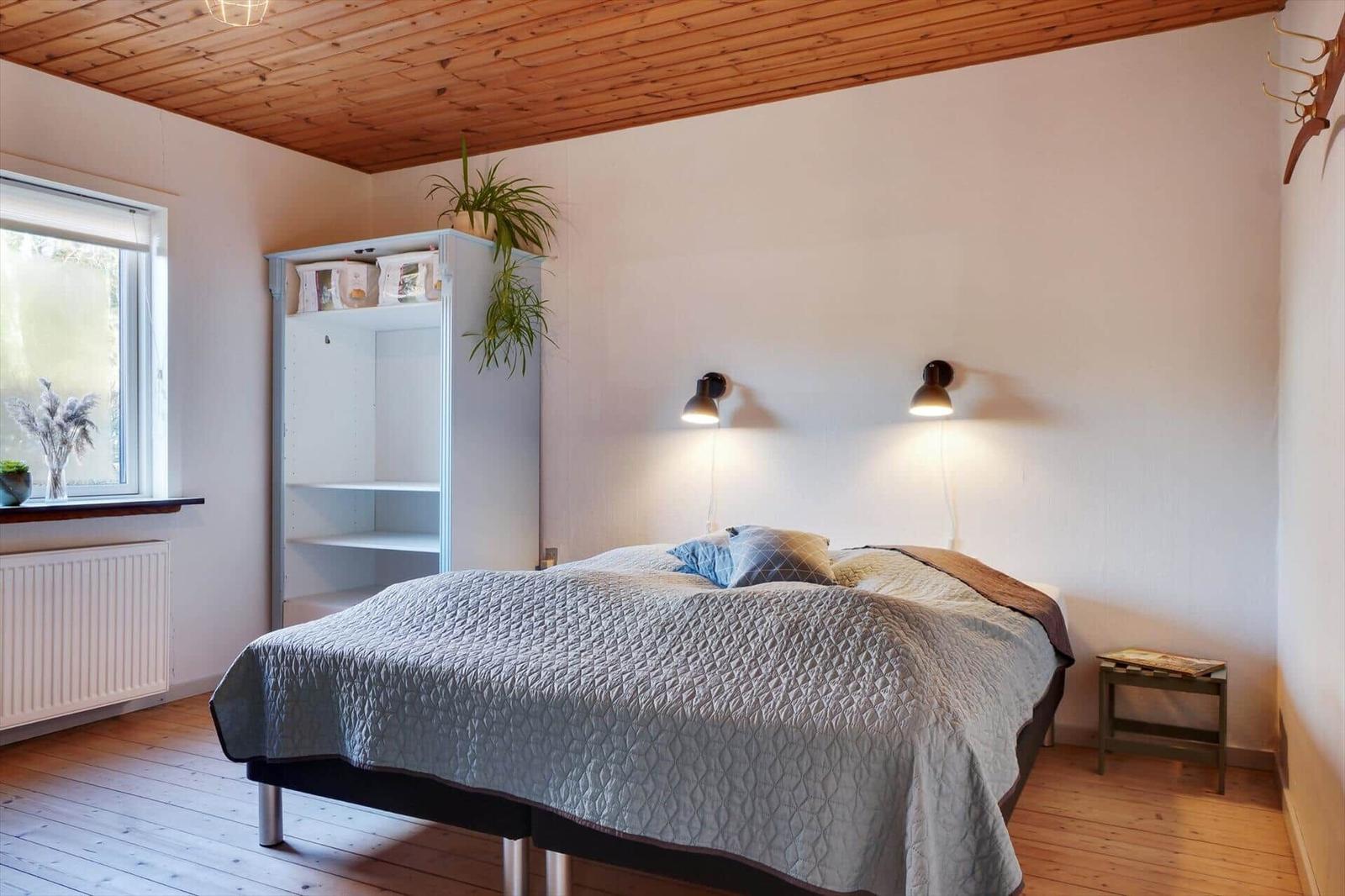 Bedroom with bed, nightstand, wardrobe, and window. Wooden ceiling and floor.