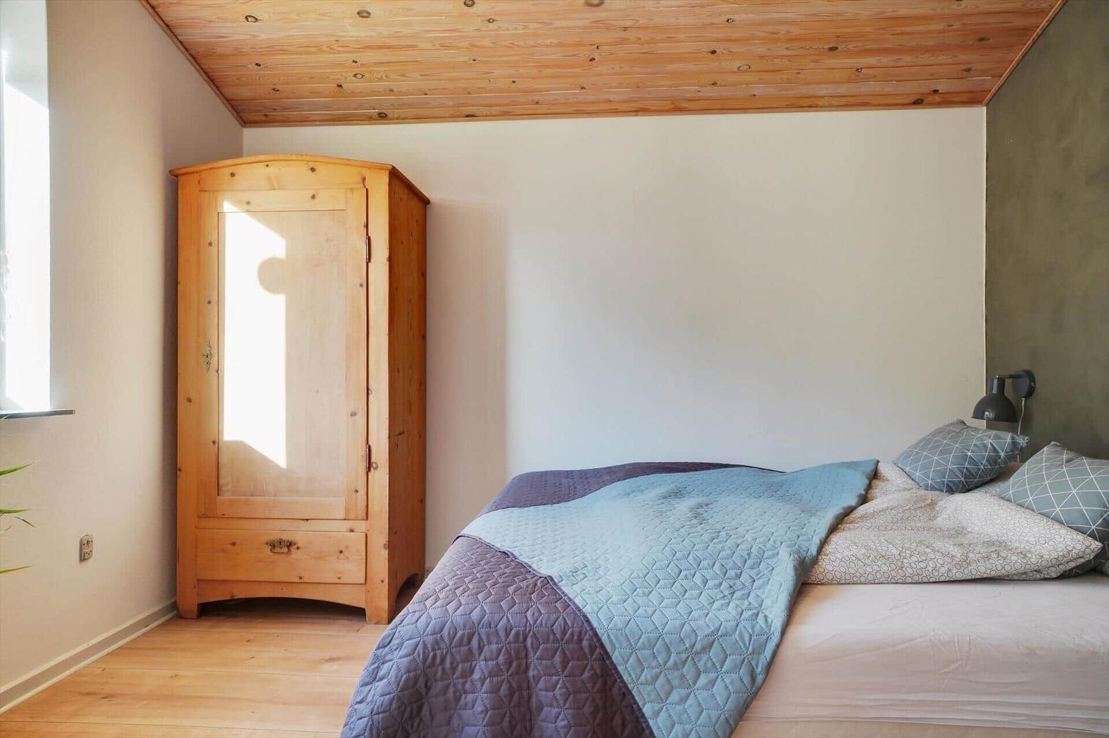 Bedroom with bed, wooden wardrobe, and wooden ceiling. Window and wall lamp visible.