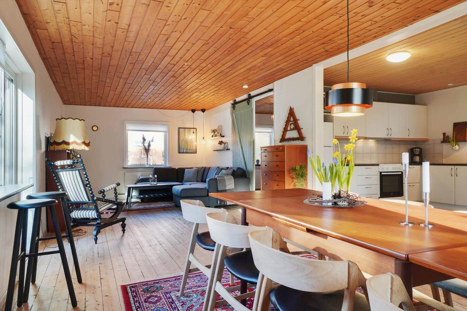 Dining area with wooden table and chairs, kitchen and living room with sofa and wooden ceiling.