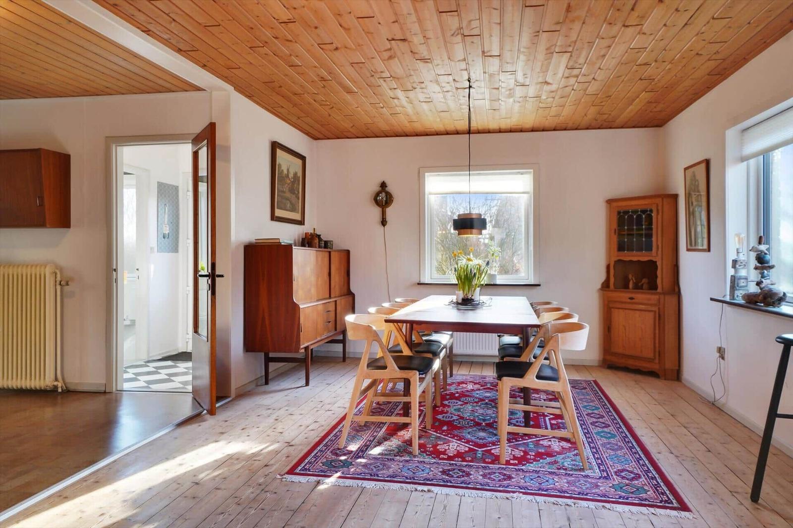 Dining room with wooden ceiling, table, chairs, and rug. Corridor to the kitchen.