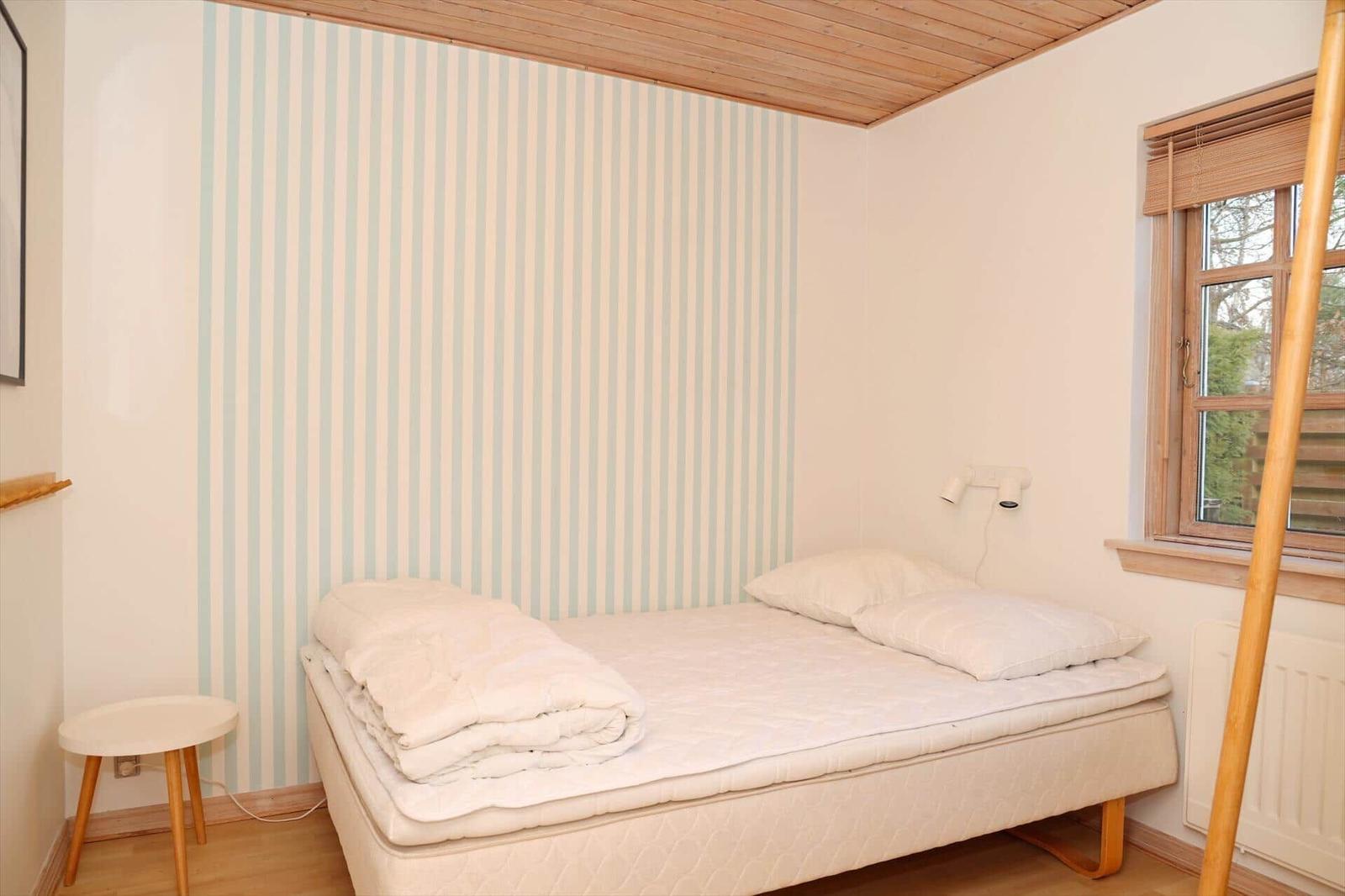 Bedroom with bed, striped wall, and wooden ceiling. Window with blinds.