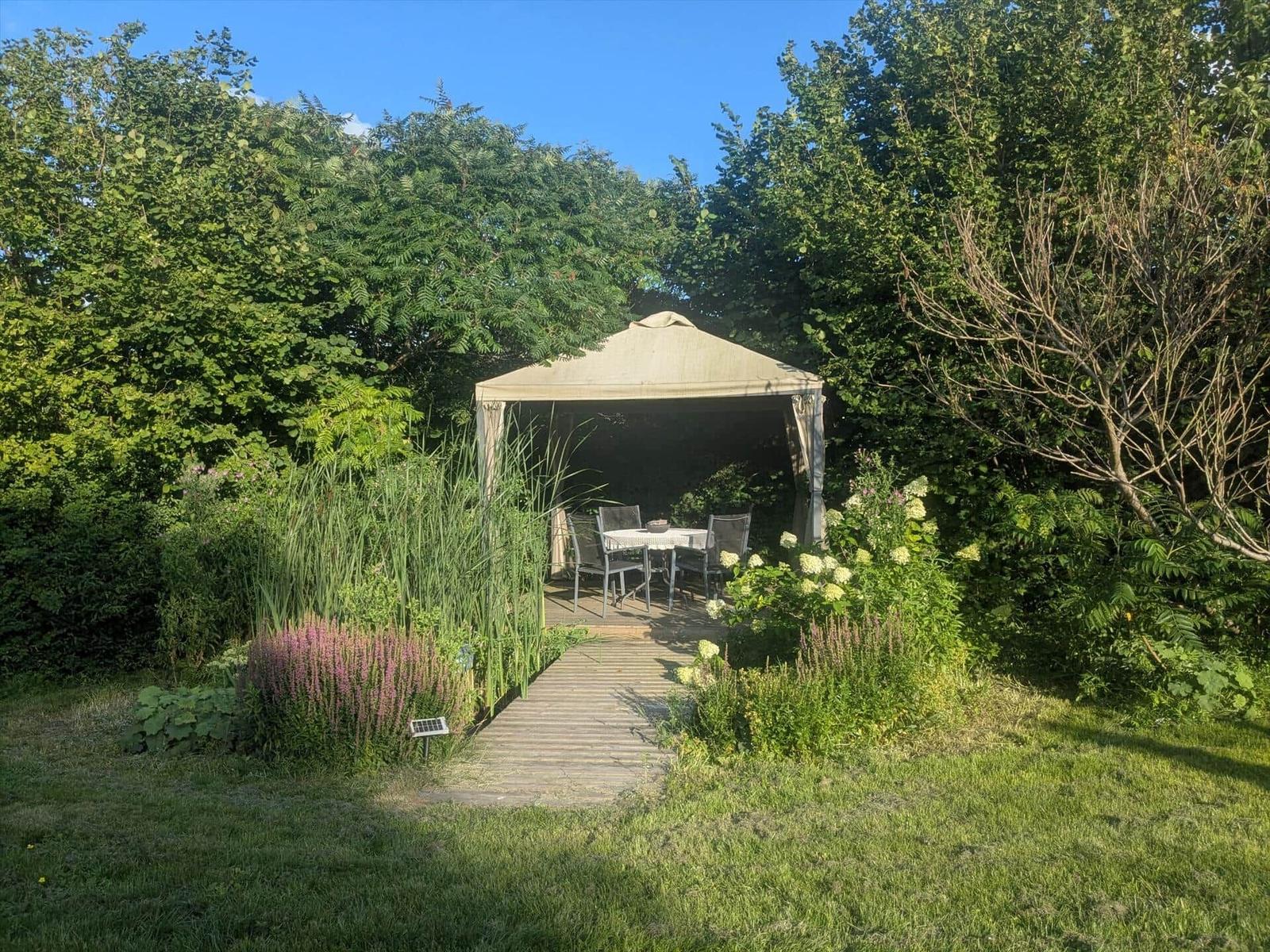 Garten mit Pavillon, Tisch und Stühlen. Holzweg führt dorthin. Umgeben von Pflanzen und Bäumen.