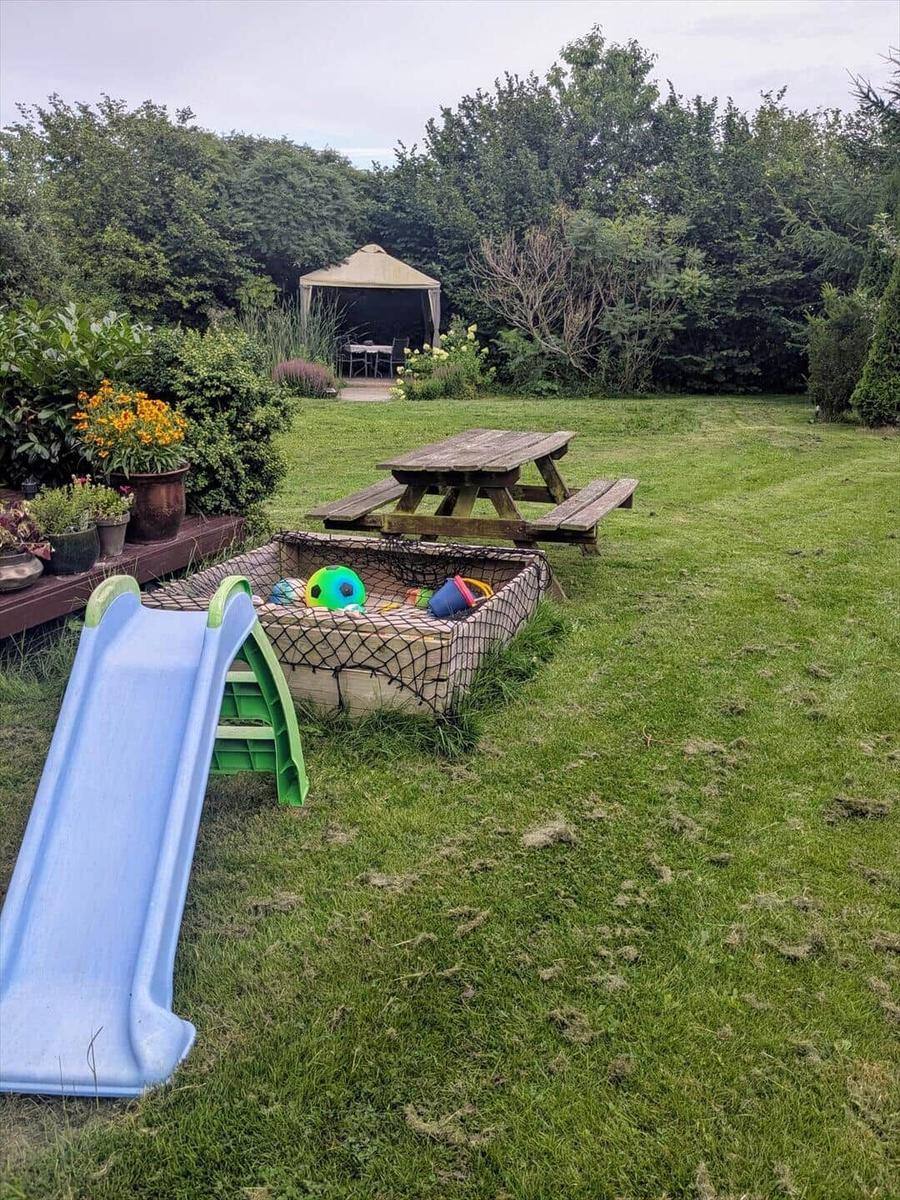 Garten mit Spielplatz, Picknicktisch und Pavillon. Grasfläche und Pflanzen sichtbar.