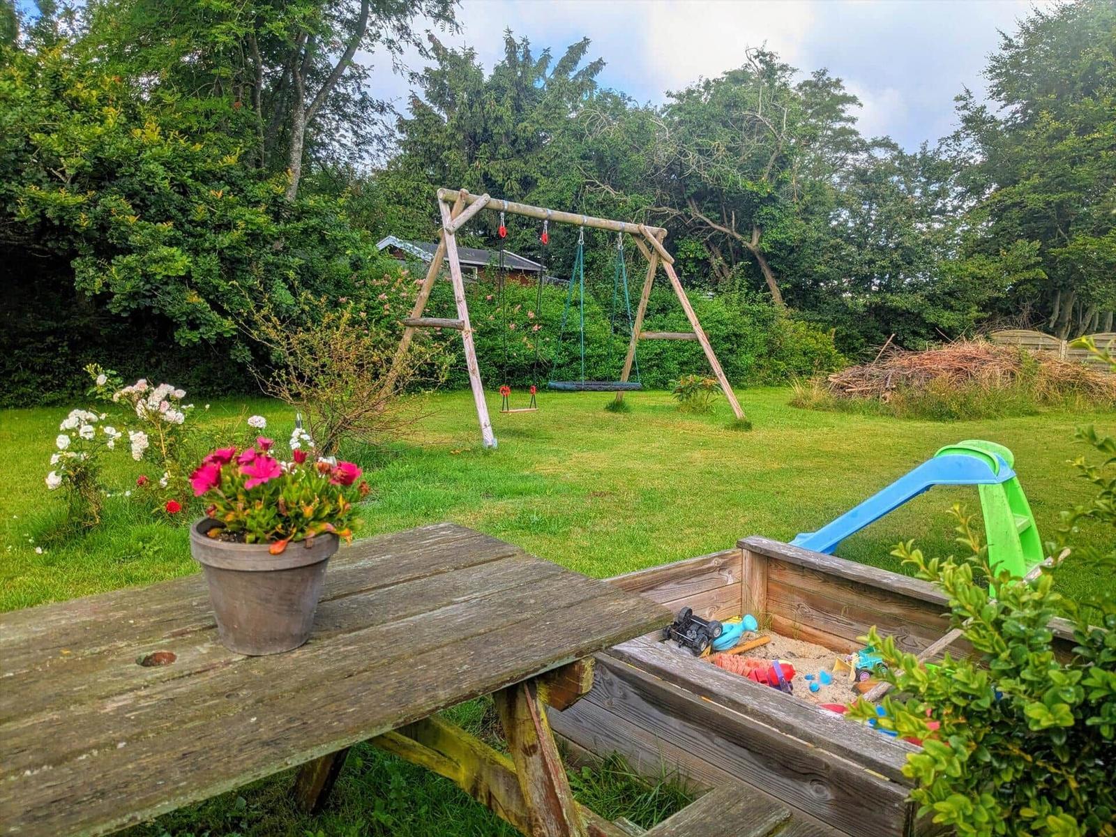 Hinterhof mit Schaukel, Sandkasten und Blumenbeet. Holztisch mit Topf im Vordergrund.