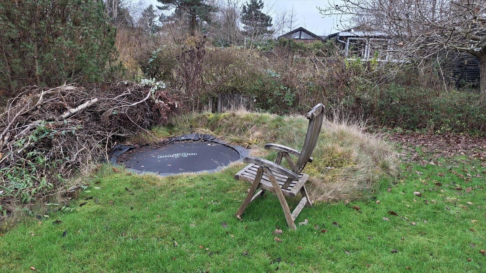 Ein Garten mit Stuhl, Trampolin und Baumstammhaufen.