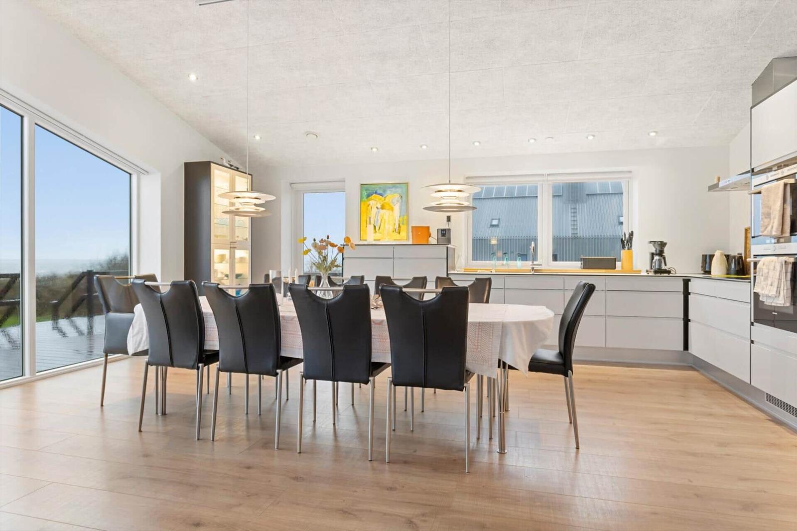 Dining room with table, chairs, and kitchen area. Large windows with view of terrace.