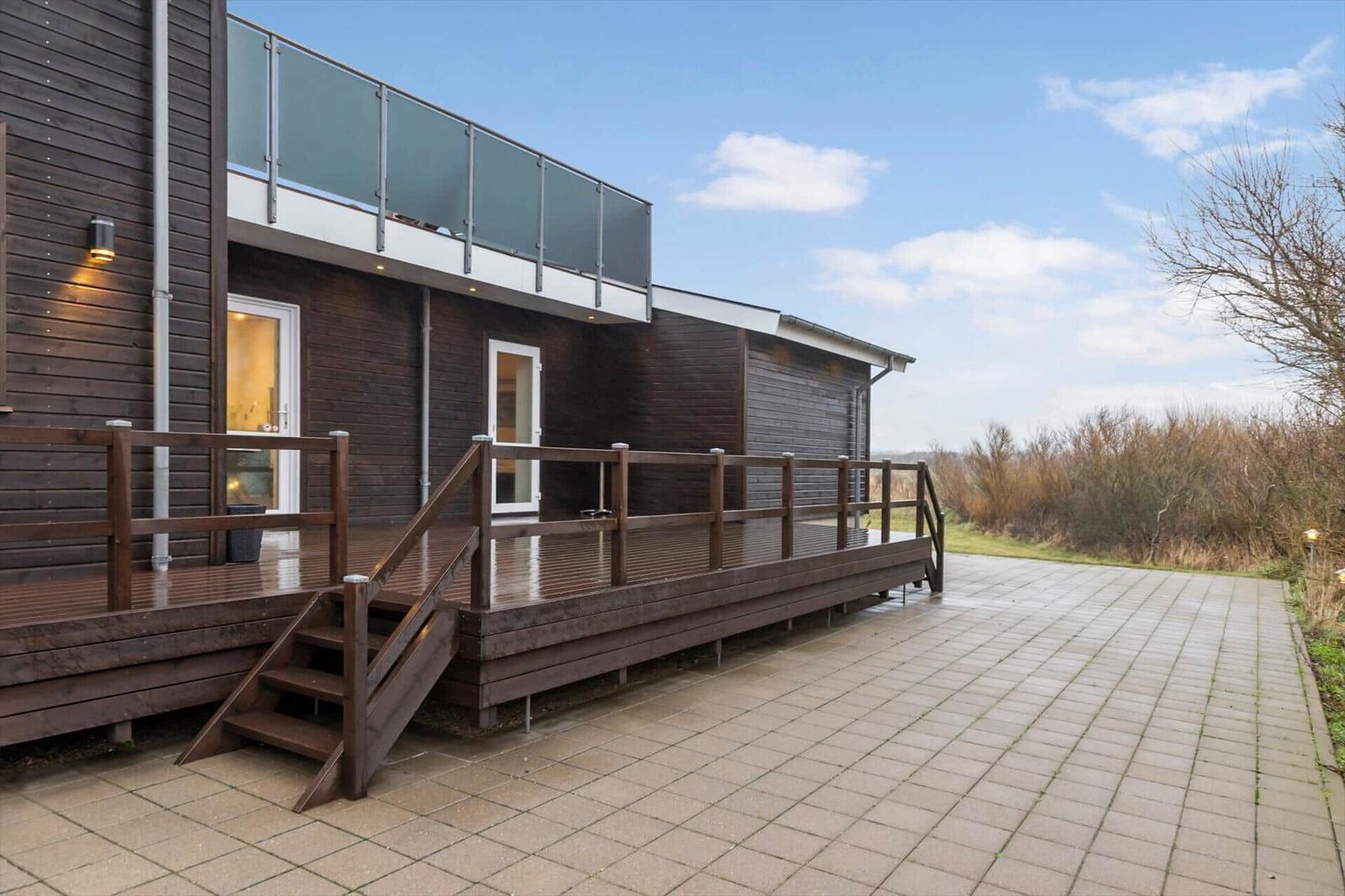 Wooden terrace with railing and outdoor stairs next to modern wooden house with glass railing.