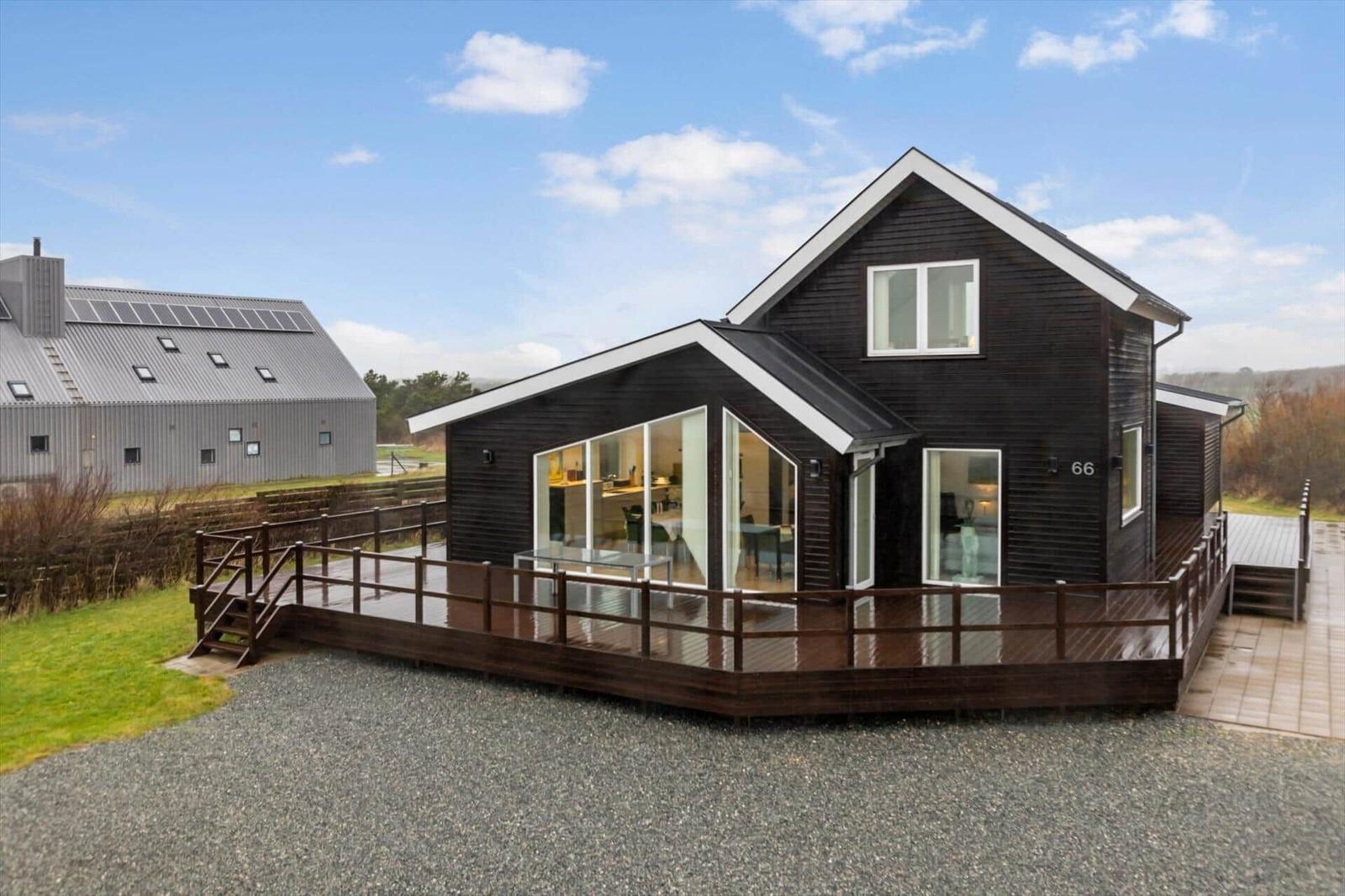 Modern wooden house with large balcony and glass front. A gray building is in the background.