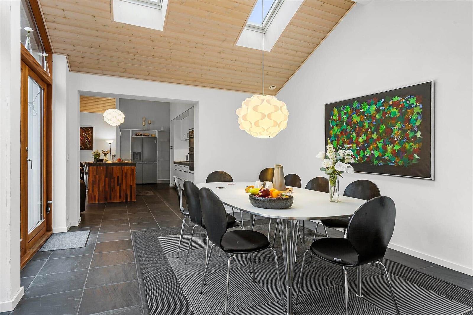 Dining area with table, chairs, and wall art. Kitchen in background. Wooden ceiling with skylights.