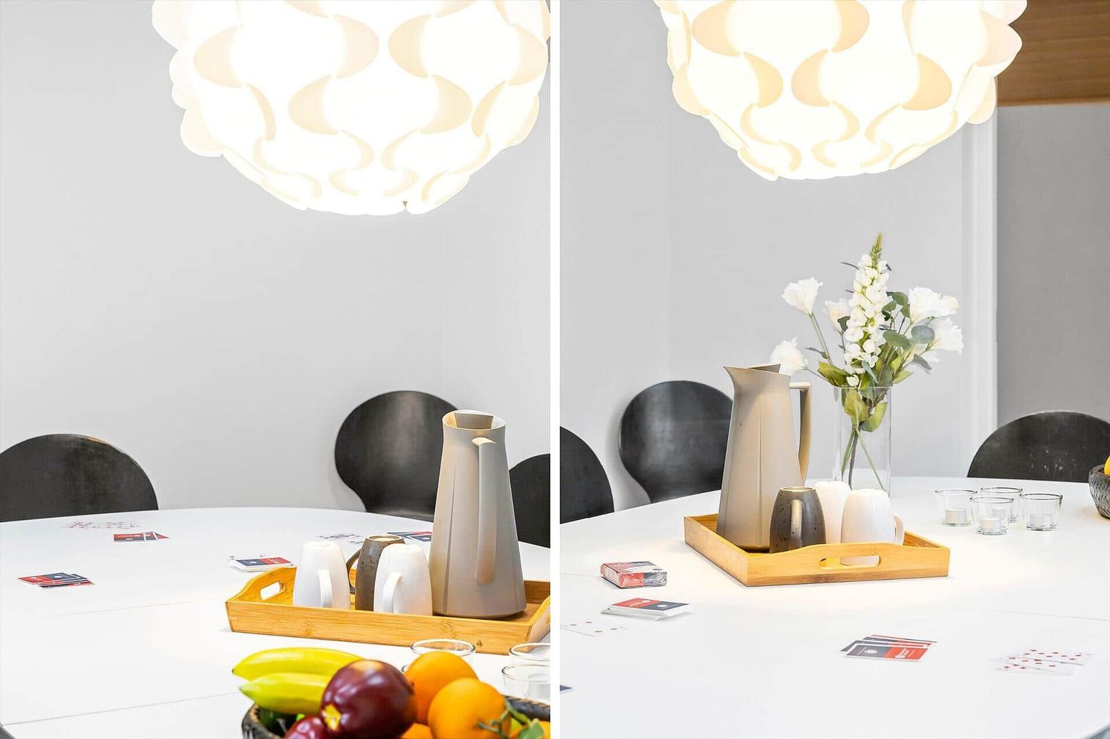 Round table with ceramic set, fruit, and flowers under a ceiling lamp.