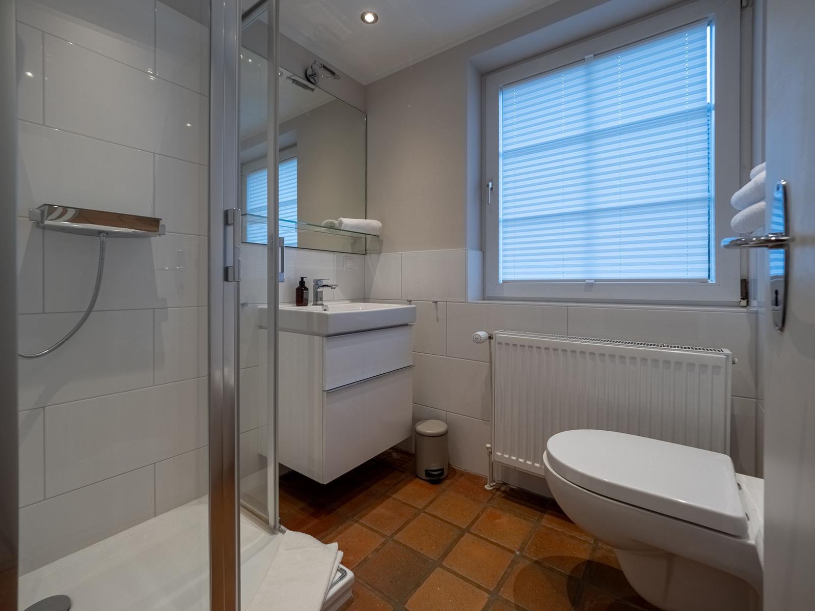 Badezimmer mit Dusche, Waschbecken und Toilette. Fenster mit Jalousie und Terrakottboden.