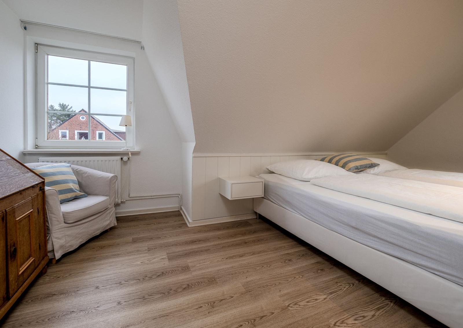 Ein Schlafzimmer mit Dachschrägen, Bett, Sessel und Fenster mit Blick auf Haus.