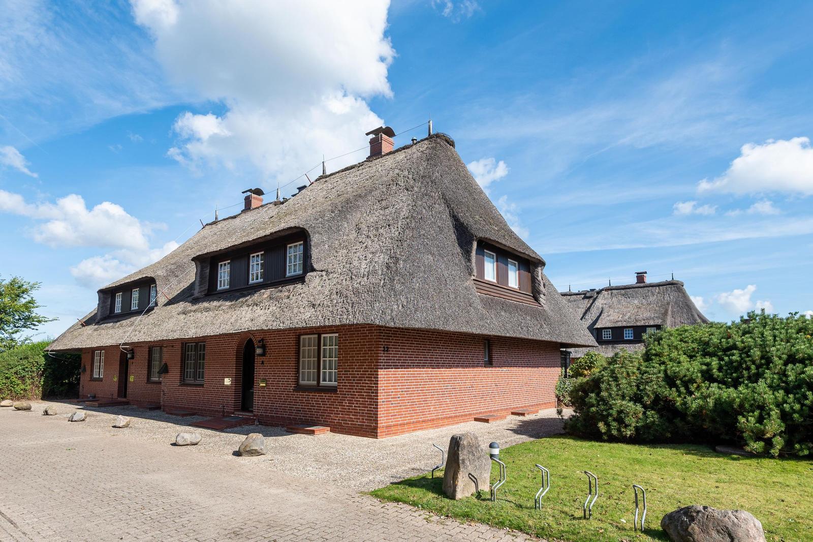 Haus mit strohgedecktem Dach, roten Ziegelmauern und grüner Umgebung unter blauem Himmel.