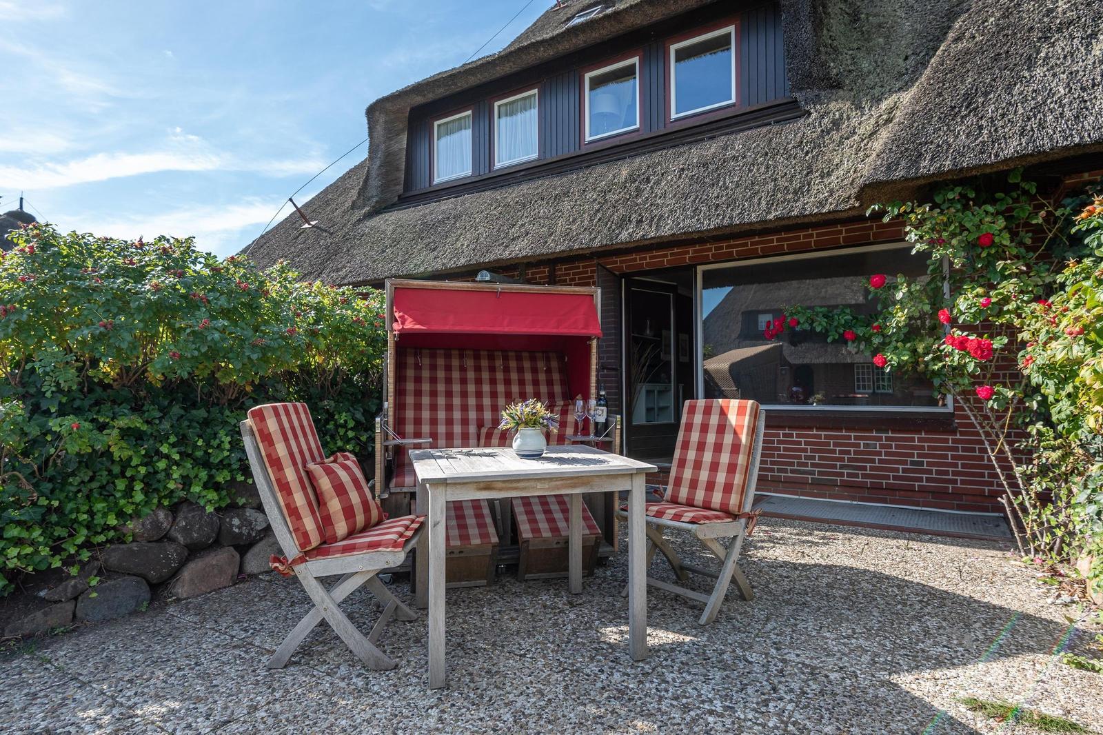 Terrasse mit Tisch, zwei Stühlen und einem Liegestuhl unter rotem Sonnenschutz.