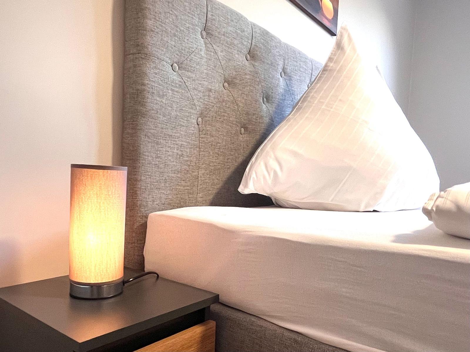 Ein Bett mit grauem Kopfteil und weißer Bettwäsche. Eine Lampe auf einem Nachttisch.