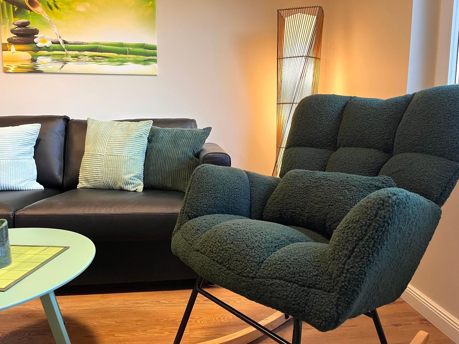 Ein dunkelgrüner Sessel steht neben einem Sofa mit Kissen. Eine Lampe und ein Bild an der Wand.