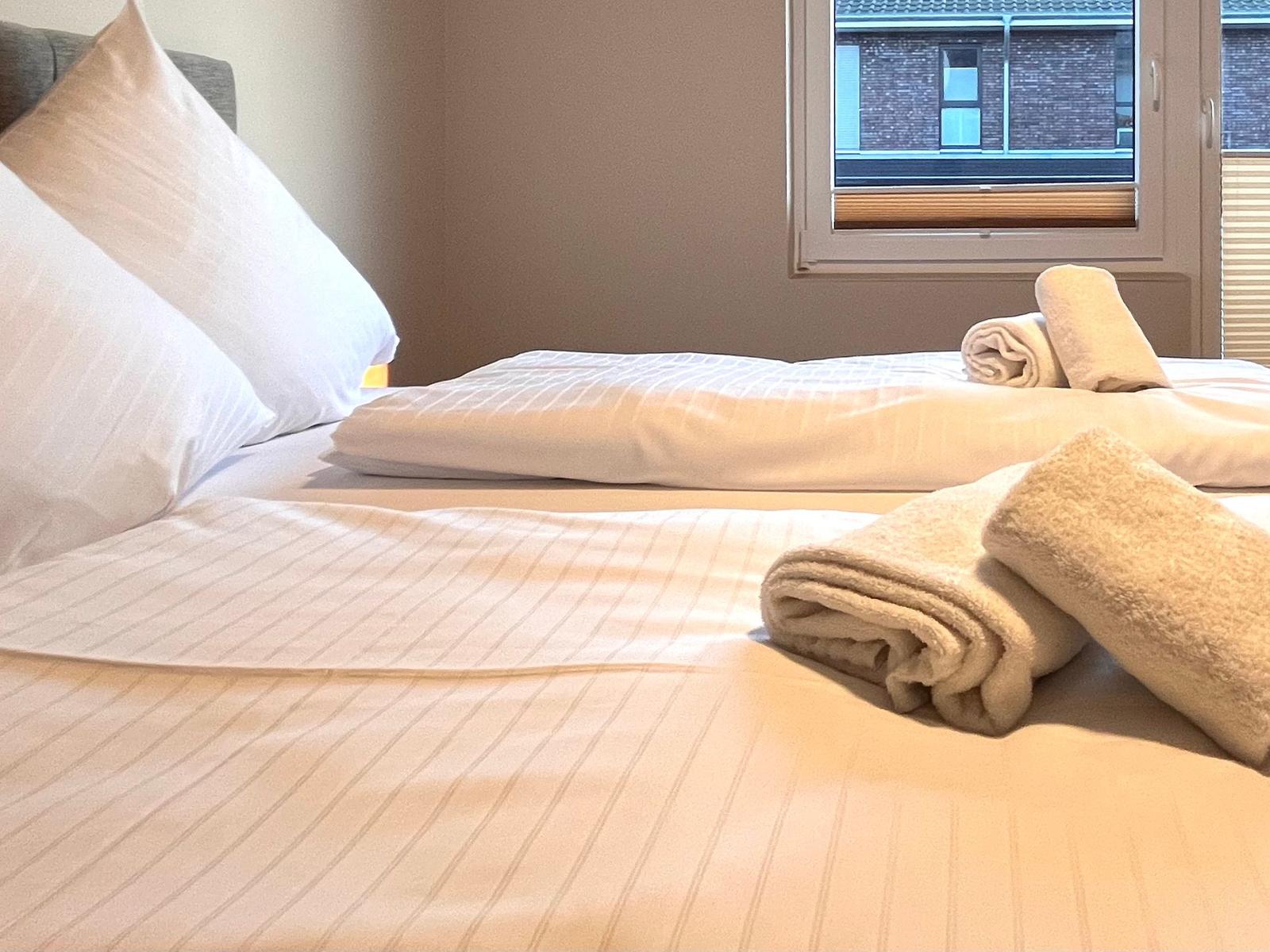 Das Bett ist mit weißen Kissen und Bettwäsche bezogen. Rollenbadetücher liegen darauf. Ein Fenster ist sichtbar.