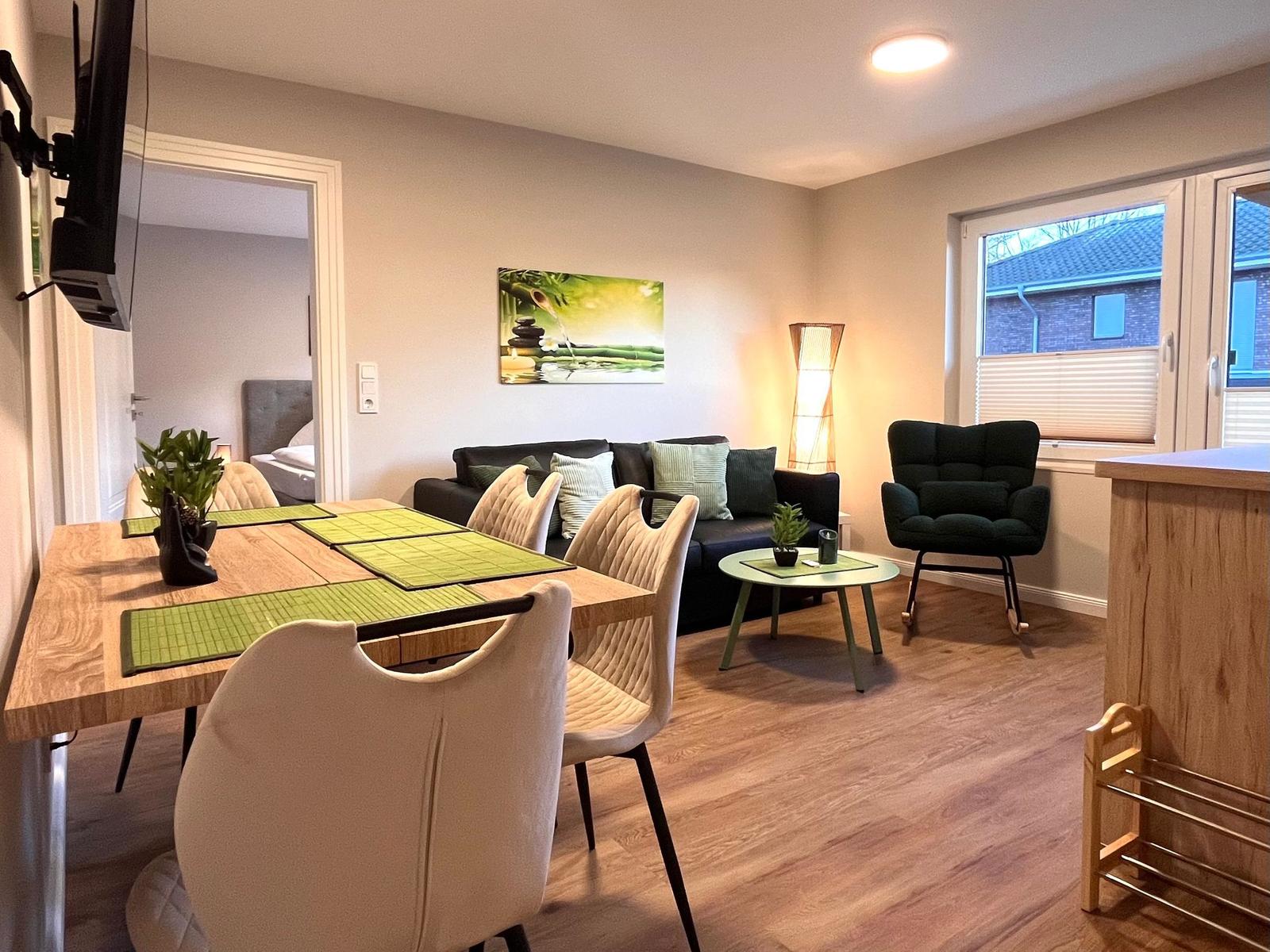 Essbereich mit Tisch und Stühlen, Wohnbereich mit Sofa und Sessel, Wandbild und Fenster.
