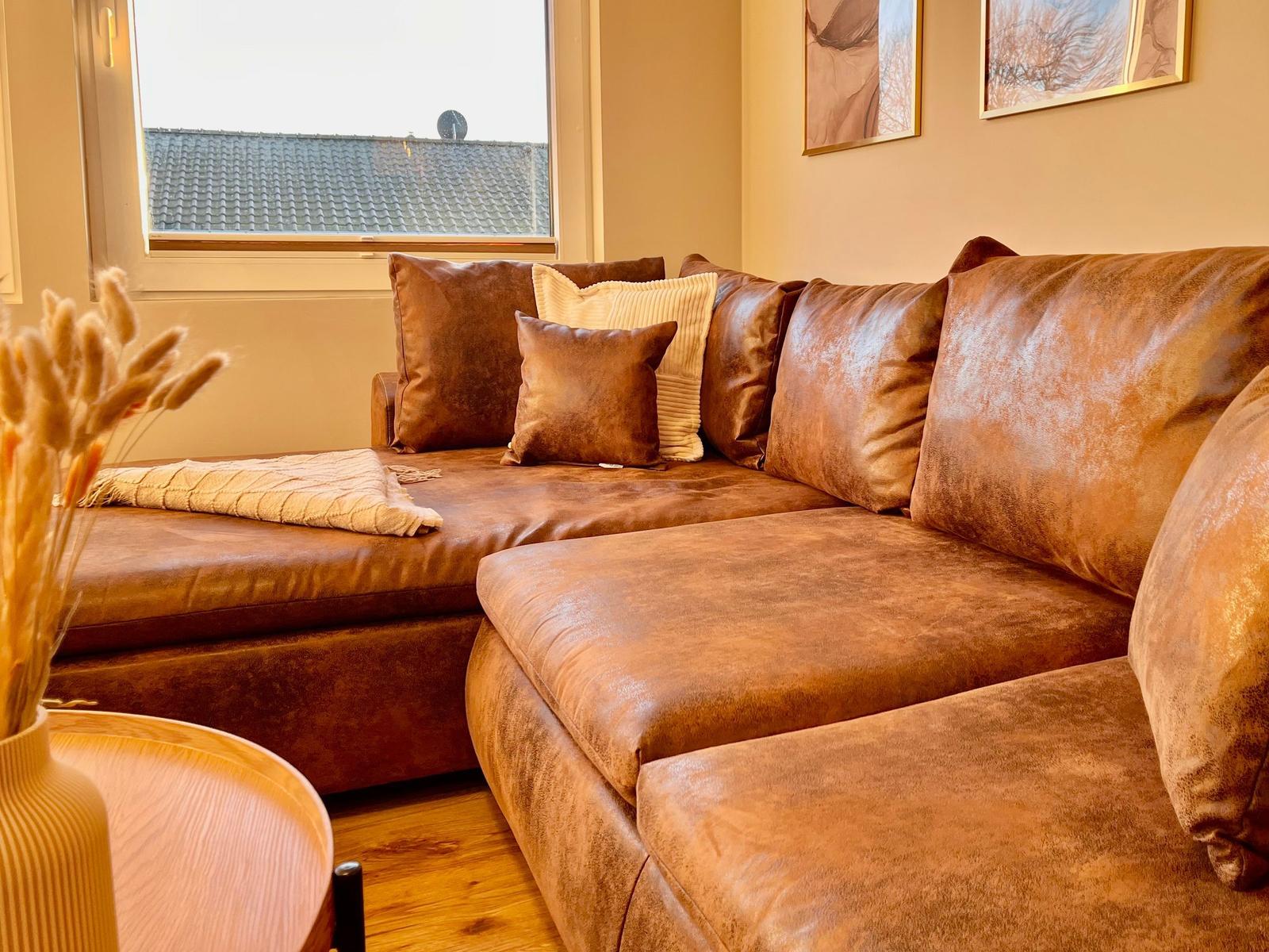 Ein braunes Ledersofa mit Kissen und einer Decke vor einem Fenster.