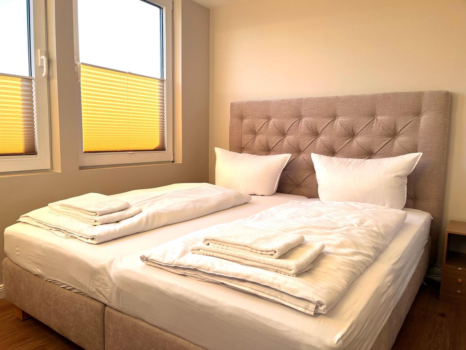 Schlafzimmer mit Doppelbett, Kopfteil und Fenster mit Jalousien.